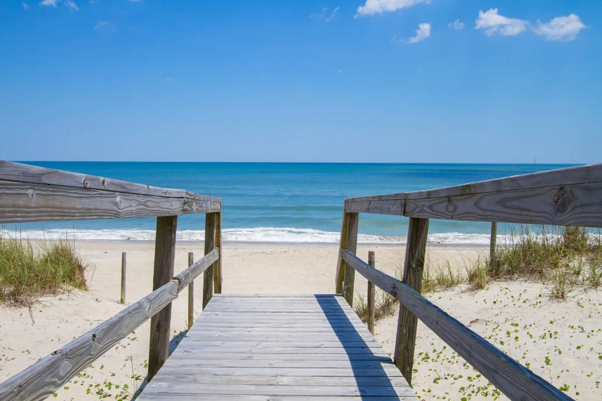 A picturesque view of a wooden walkway stretching across soft sands to the ocean, framed by a clear blue sky. The scene invokes a sense of arrival, inviting guests to step onto the beach and enjoy the waves, sand, and sun.