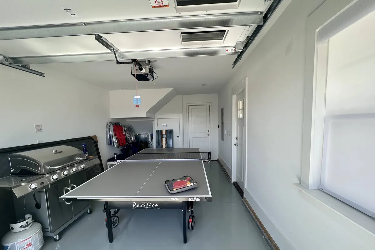 This image captures a dynamic garage setup featuring a ping pong table at the center, a premium gas grill positioned along one wall, and ample storage space for various equipment. The scene conveys a lively, playful atmosphere ideal for friendly competitions, casual cookouts, and preparing for outdoor adventures.