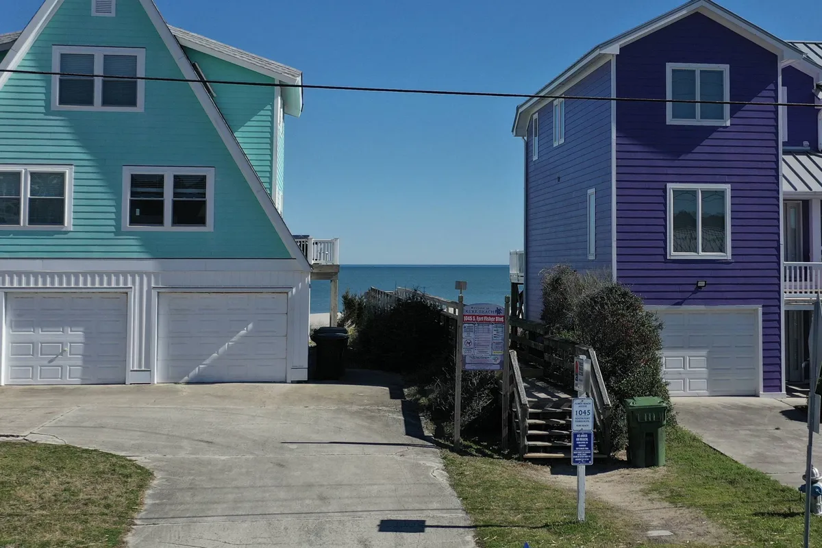 The image features two colorful beach houses with a direct view of the ocean and a sandy pathway leading to the beach. The cheerful mood and visible proximity to the shoreline capture the essence of family outings and easy access to beachside fun.