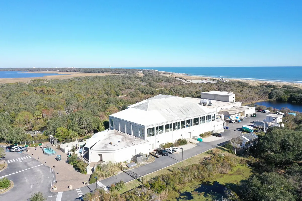 This image shows a large building surrounded by lush greenery, with an expansive view of the ocean and a parking area in the foreground. The scene is inviting and highlights both the natural landscape and a community-focused space, hinting at local attractions and opportunities for outdoor exploration.