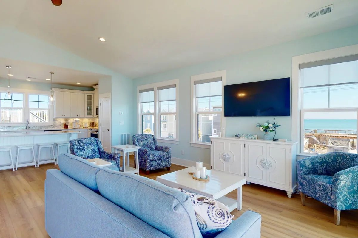 A bright interior combining a comfortable sofa, a dedicated dining area, and expansive windows offering direct ocean views. The presence of a TV and tasteful decor underscore the room's purpose as a versatile gathering spot for relaxing, dining, and entertaining.