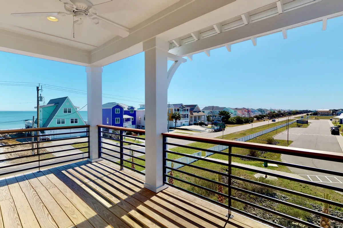 The image showcases the exterior of a luxury townhome at Kure Beach, with a spacious balcony overlooking the ocean, vibrant neighboring houses, and a picturesque street below. The scene suggests relaxation and togetherness, with opportunities for sunset dinners, morning coffee, and easy beach access just steps away.