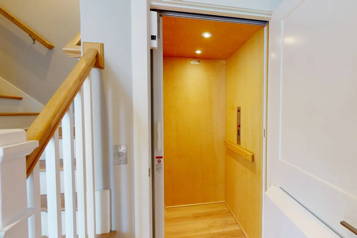 The interior of a private elevator, featuring a wooden handrail and a glimpse of a staircase. The inviting design emphasizes accessibility and the exclusive nature of the upper-level entry.