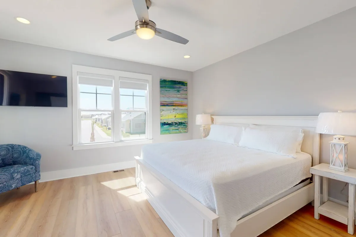 A serene queen bedroom with a comfortable bed, modest yet elegant artwork, a reading chair, and gentle natural light streaming through a window. The decor is simple and soothing, aligned with a restful, private retreat.