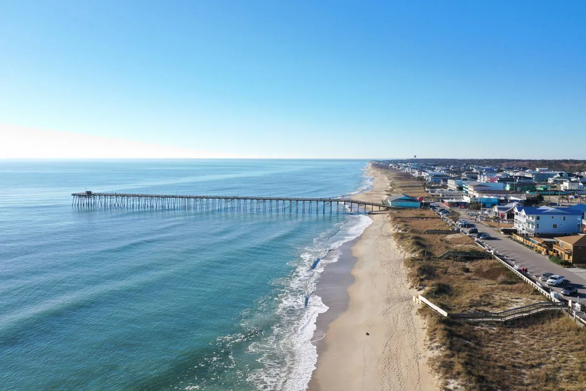 This image showcases the iconic Kure Beach Pier extending over the ocean, with the beach, waves, and nearby houses in the background. The scene hints at both fishing adventures and leisurely walks along the shore, capturing the essence of the local beach culture.