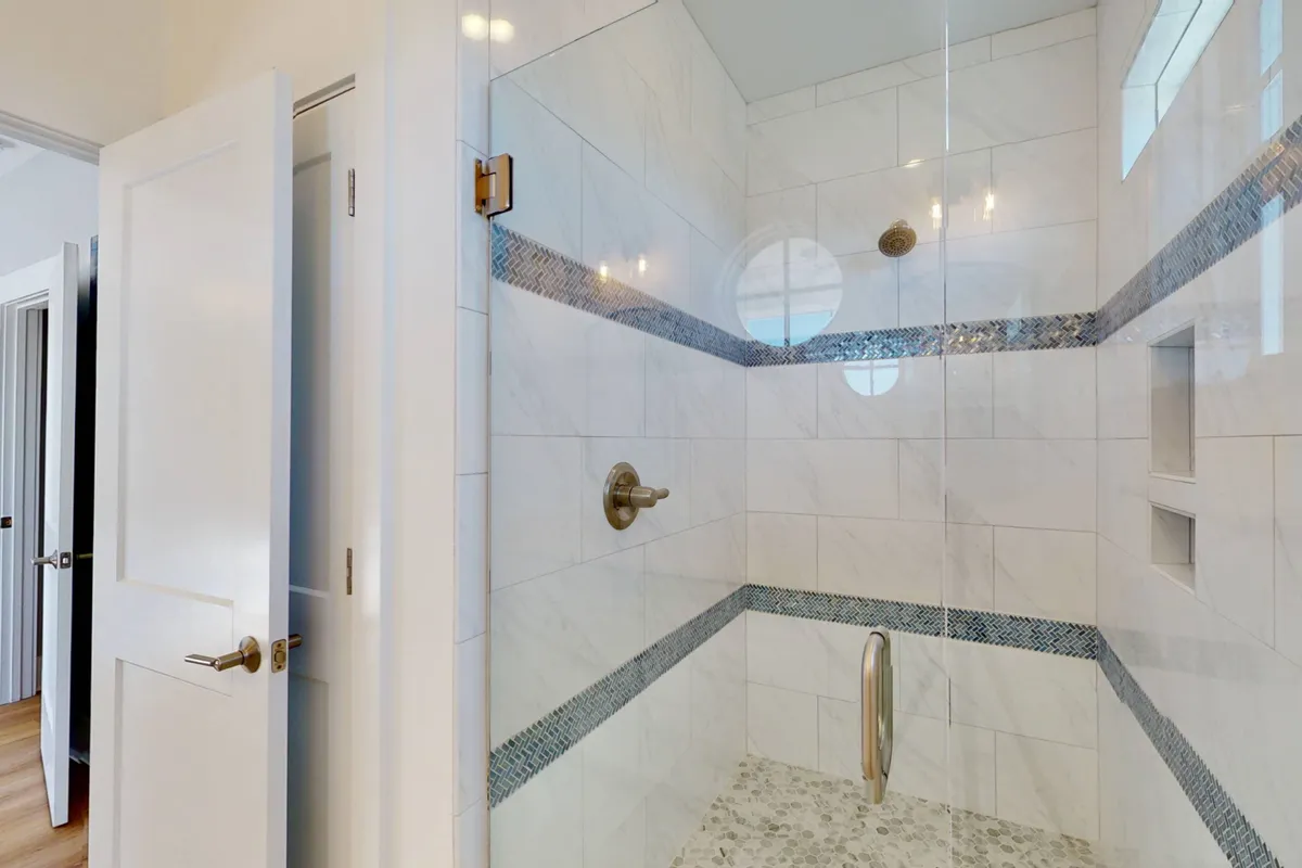 A luxurious, spa-inspired bathroom featuring a glass-enclosed shower, marble tiles, a decorative mosaic strip, and stylish fixtures, all illuminated by soft, serene lighting. The atmosphere evokes relaxation and modern comfort.