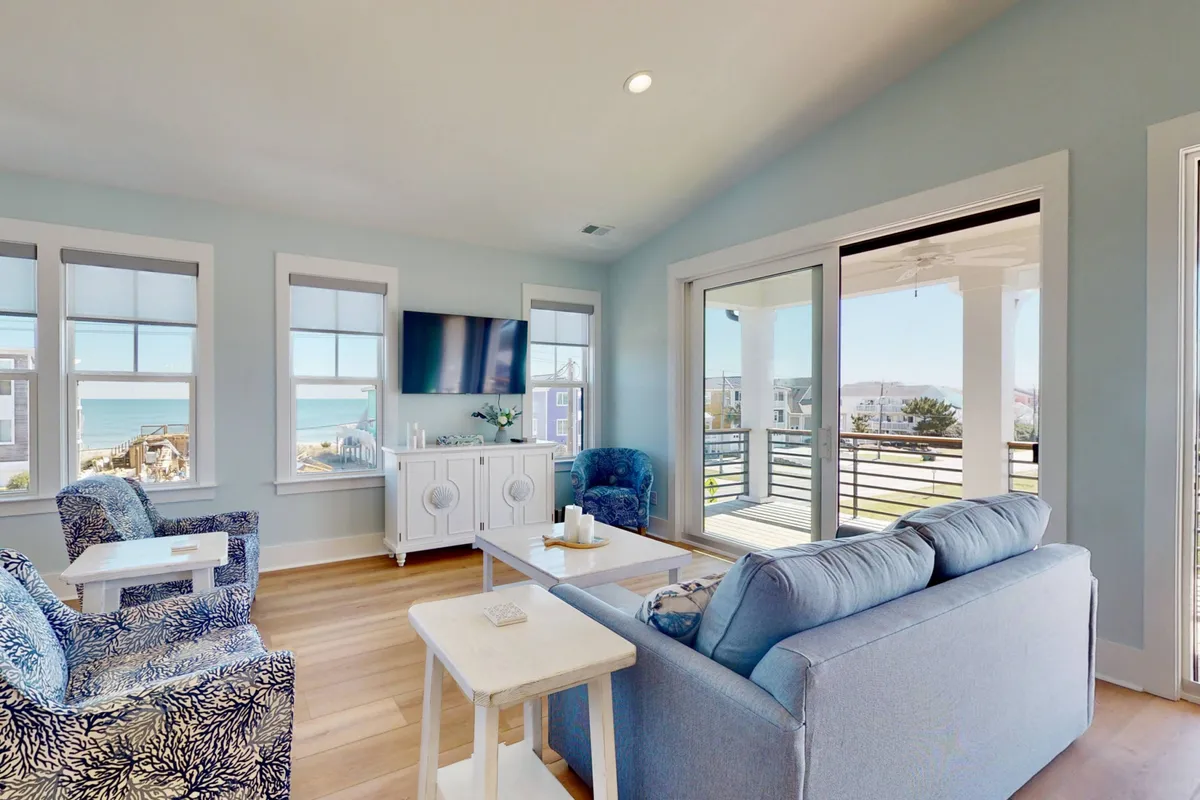 A spacious living area featuring large windows framing a breathtaking ocean view. The room is furnished with a blue sofa, decorative chairs, a coffee table, and a television, all bathed in natural light from the windows. The setup invites both relaxation and social gatherings, with designer touches enhancing the calm, serene mood.