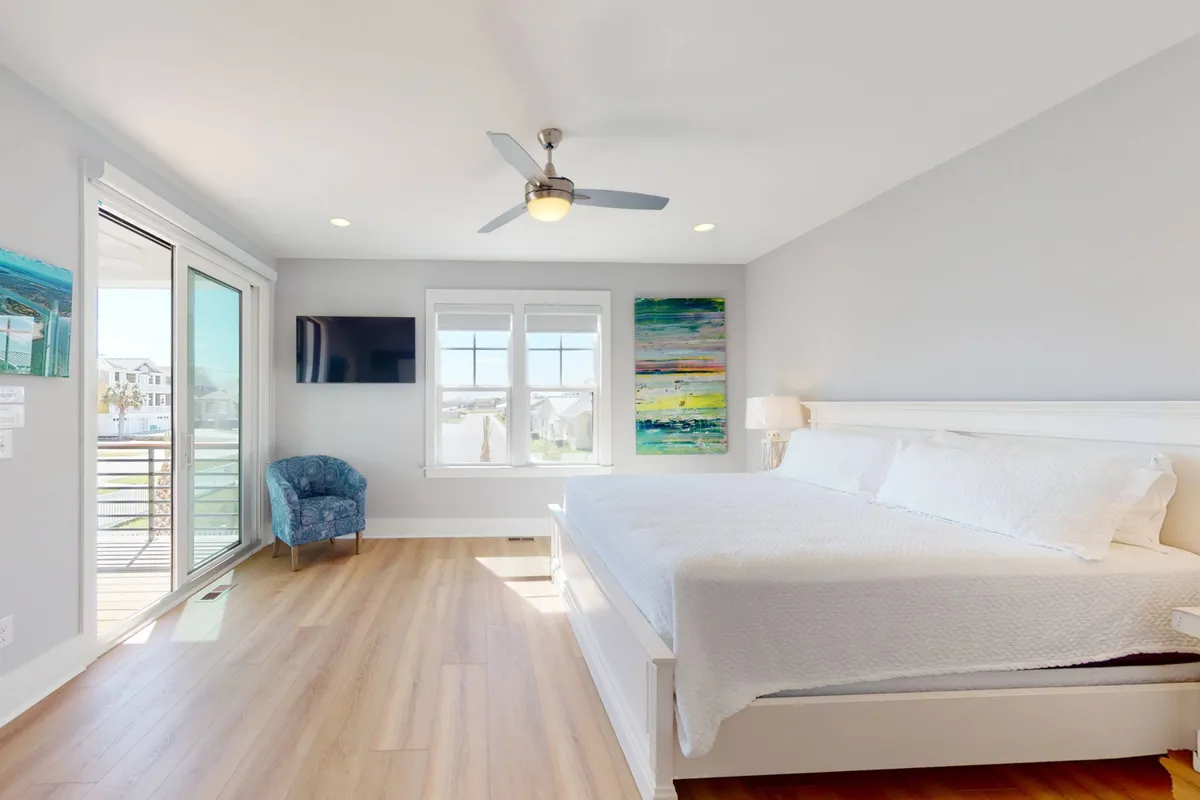 A spacious bedroom with a large bed adorned with plush bedding and fresh linens, highlighted by calming artwork on the wall and a comfortable blue chair positioned near windows that flood the room with natural light. The space exudes tranquility and privacy, ideal for unwinding after a day at the beach.